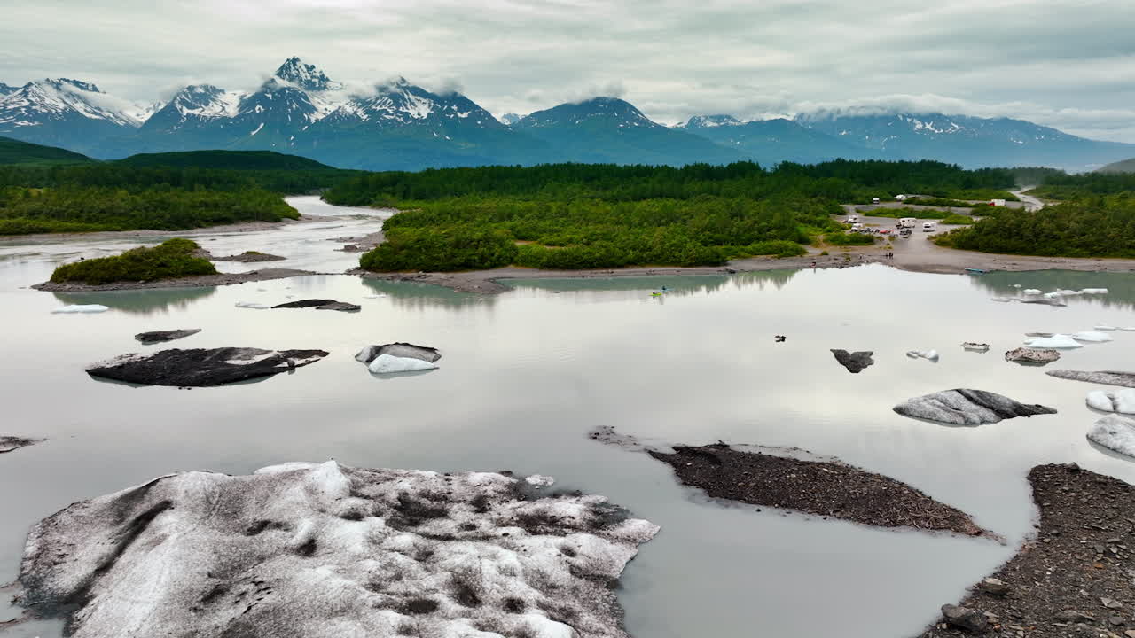 Footage above the river with melting ice boulders. Campers are at the waterfront on the other side. Mountain range covered with clouds at tops at backdrop. Alaska, USA
