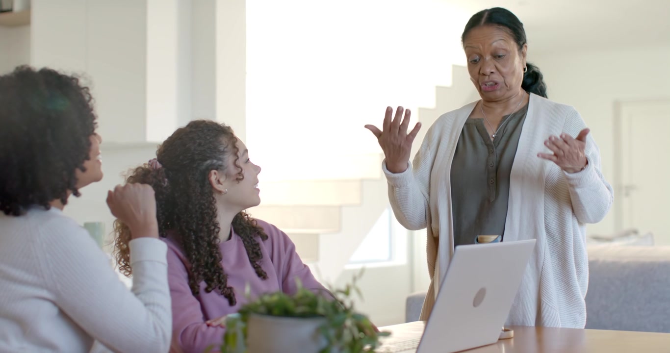 Senior African American woman walking to table, leaning on laptop and explaining to women