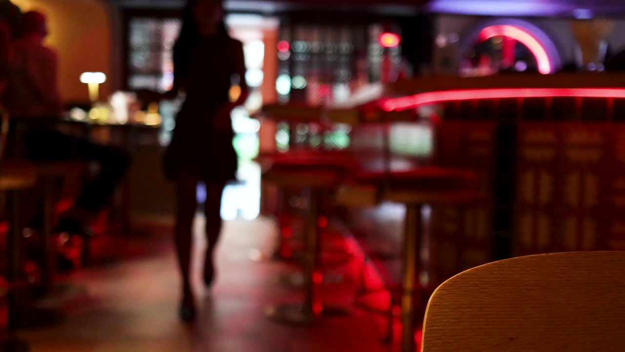 A silhouetted woman enters a contemporary bar with red neon accents, captured in soft focus with moody, low ambient lighting and a stationary camera