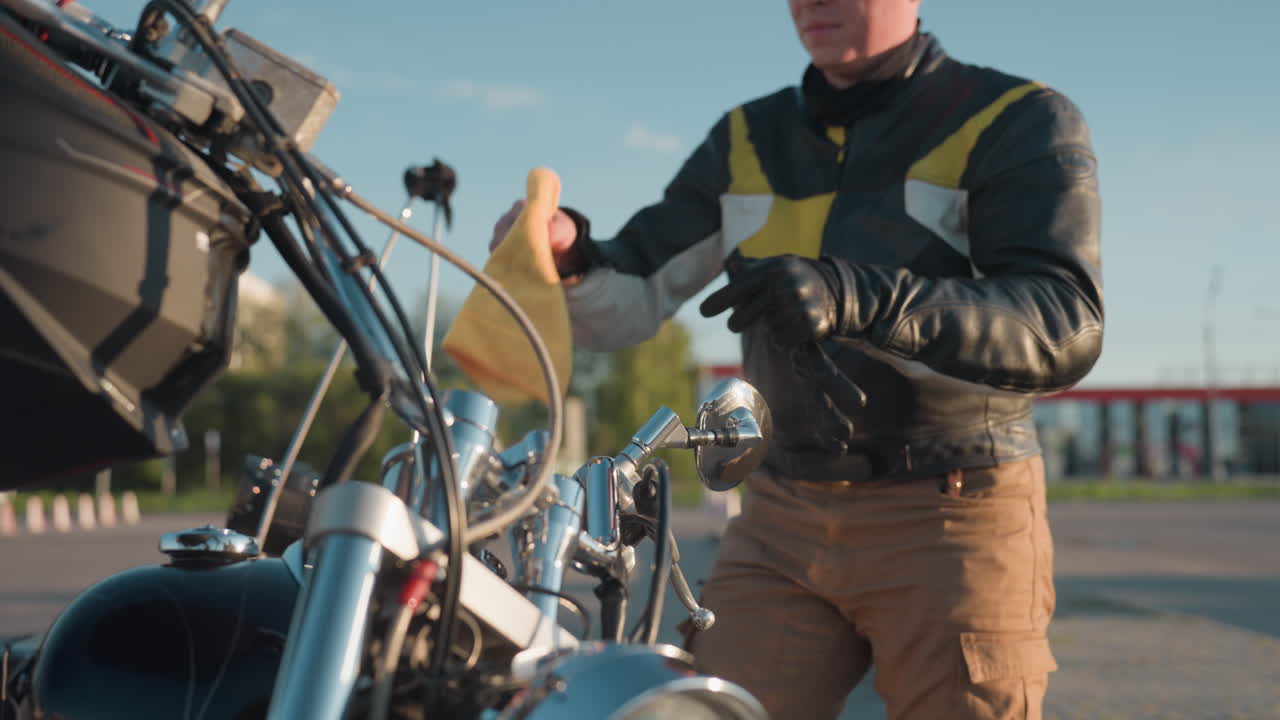 Motorcycle rider wearing leather jacket carefully cleaning bike with rag under clear sky, sunlight reflecting on chrome mirror while standing in open outdoor space with blurred greenery in background