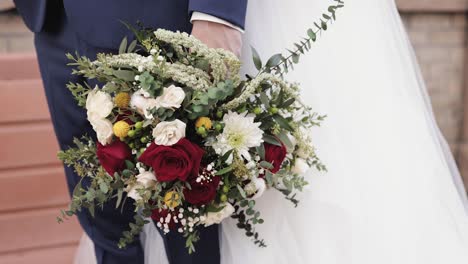 Braut-Und-Bräutigam-Im-Freien-Umarmen-Sich-In-Hochzeitskleidung-Und-Halten-Einen-Orangefarbenen,-Rosa-Und-Weißen-Blumenstrauß-In-Ihren-Händen-1080p-60fps