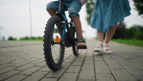 leg view of a child riding a blue bicycle while a woman in a blue dress follows closely from behind