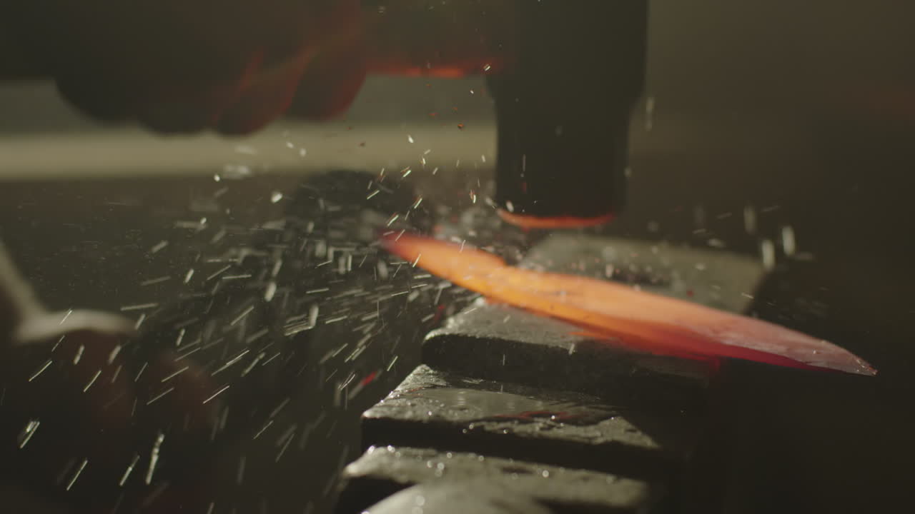 A blacksmith hits heated metal with a wet hammer causing water to spray around the anvil
