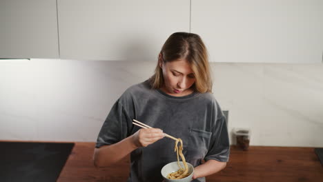 overhead view of young woman in gray casual outfit eating breakfast noodles with chopsticks, focused on meal, standing in bright modern kitchen with white walls and wooden countertop