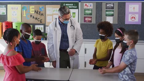 Diverse-schoolteacher-and-schoolchildren-standing-disinfecting-hands,-all-wearing-face-masks