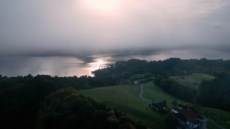 An-aerial-view-of-a-small,-rectangular-house-with-a-red-roof-and-a-wooden-dock,-located-on-the-edge-of-a-calm,-blue-lake-surrounded-by-lush-green-trees-under-a-partly-cloudy-sky