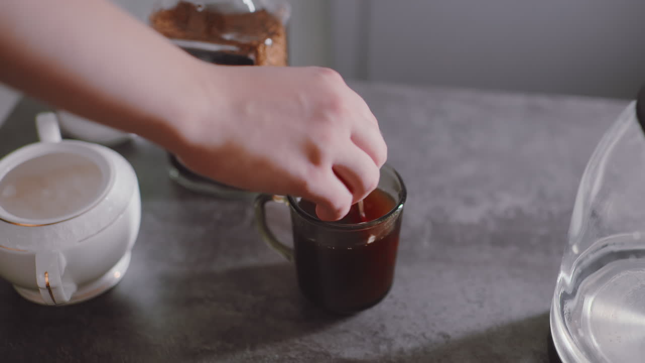 Close up of hand scooping sugar from white container to add into coffee cup beside jar of granules, capturing detail of sweetener preparation during warm drink making on kitchen surface
