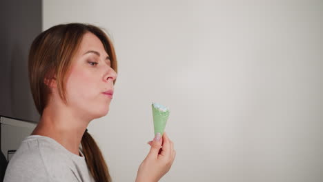 close up of young woman with brown hair wearing grey t-shirt gently savoring colorful ice cream cone, standing relaxed against modern fridge in bright minimalistic kitchen