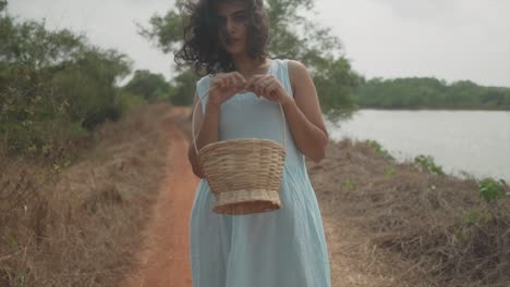 slow motion handheld shot of a young charming indian woman in a light blue dress with a wooden basket in her hands standing in front of a lake in nature on a dried footpath looking into the camera