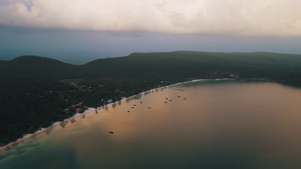 Beautiful reflection of the clouds in the streched bay of Koh Rong Sanloem where multiple boats are moored close to the white sandy beach with high green palm trees. High angle drone panning shot