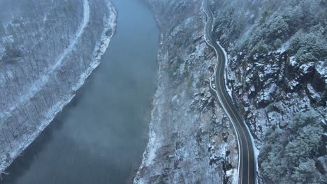 Imágenes-Aéreas-De-Un-Desvío-Panorámico-Y-Nevado,-Un-Sinuoso-Camino-Del-Valle-De-La-Montaña-Durante-Una-Tormenta-De-Nieve-Con-Pinos,-Un-Río,-Una-Carretera-De-Montaña,-Acantilados-Rocosos-Y-Bosques-Durante-El-Invierno-En-Un-Día-Frío-Y-Azul