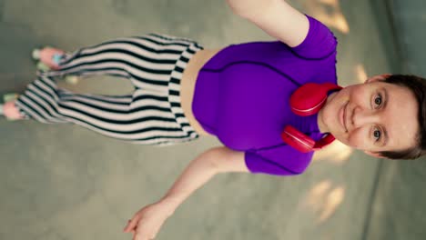 Vertical-video:-A-girl-in-a-purple-top-red-headphones-in-striped-pants-and-pink-roller-skates-rides-in-a-skate-park-and-takes-a-selfie-showing-V-sign-in-a-skate-park-in-summer