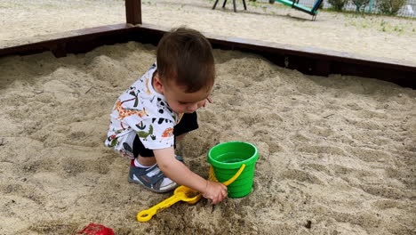 dark-haired baby boy sitting in the sandbox puts the sand into his bucket. smiley toddler washes hands in the bucket.