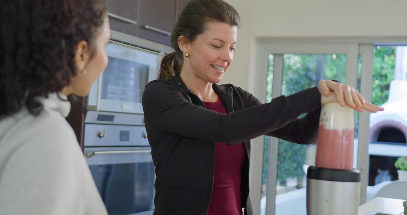Due donne adulte in cucina, una donna posiziona il boccale del frullatore sulla base, stabilizzandolo e frullando una bevanda.