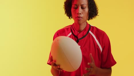 Portrait-of-african-american-female-rugby-player-with-rugby-ball-over-neon-yellow-lighting