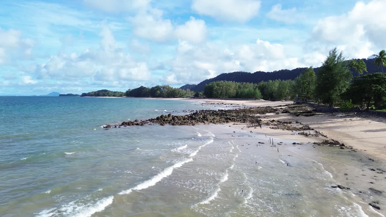 Rekaman aerial drone dari garis pantai berbatu di Long Beach Koh Lanta, Thailand, menampilkan pohon palem yang rimbun di latar depan dan bungalow tropis di sepanjang pantai, dengan perairan berwarna pirus pada hari berawan