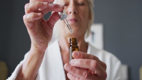 happy senior caucasian woman sitting on bed in bedroom, applying serum