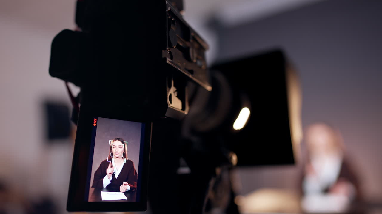 Camera display showing a female reporter speaking to camera. Close up. Studio footage of blog or program. Blurred backdrop.
