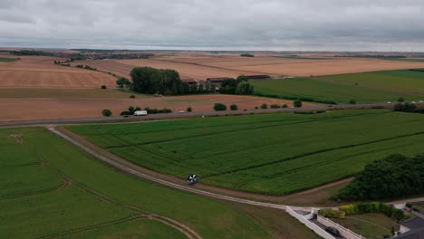aerial drone follows a farm vehicle driving on a dirt track between green agricultural fields in the french countryside, with a highway in the background. overcast day