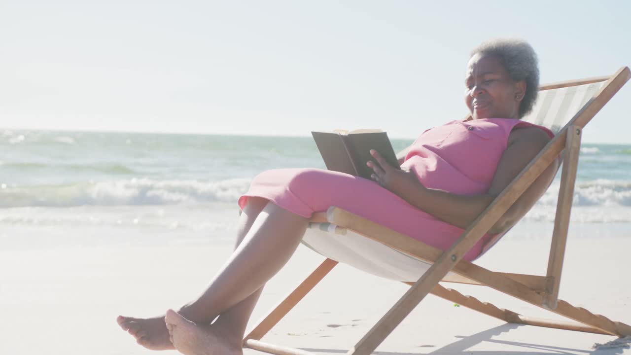 feliz mujer afroamericana mayor sentada en una silla de cubierta y leyendo un libro en la playa, en cámara lenta
