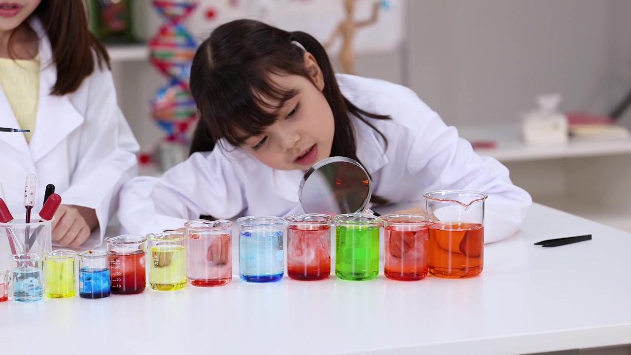 Kaksi tyttöä laboratoriovaatteissa tutkii värikkäitä kemiallisia liuoksia suurennuslasilla luokkahuoneessa.