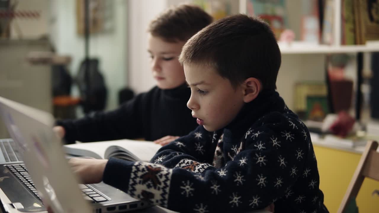 замедленные кадры двух маленьких мальчиков в теплых уютных свитерах, сидящих в классе программирования перед ноутбуками, пытающихся научиться пользоваться этими гаджетами.