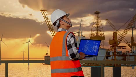back view of asian male engineer with safety helmet working on laptop with wind turbine blueprint with offshore hydrogen production