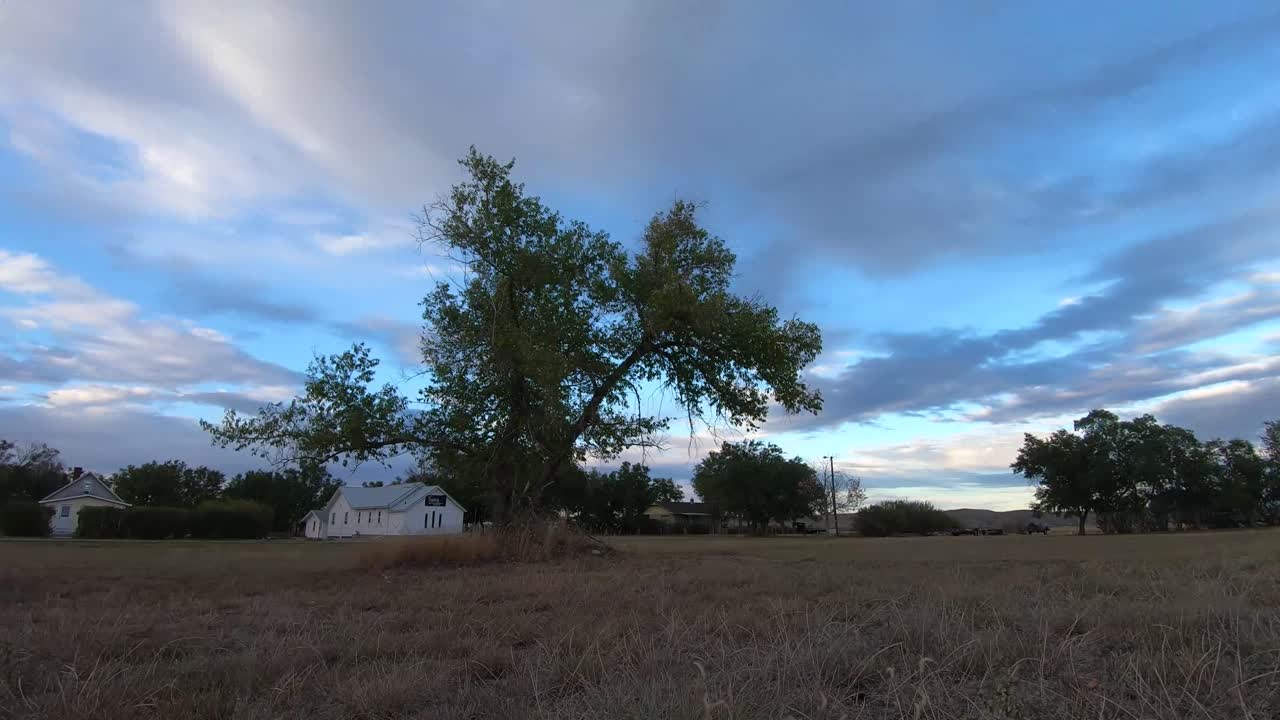 타임랩스 - 낮 동안 구름이 머리 위로 빠르게 움직이는 배경에 교회가 있는 들판 한가운데에 있는 큰 나무 하나