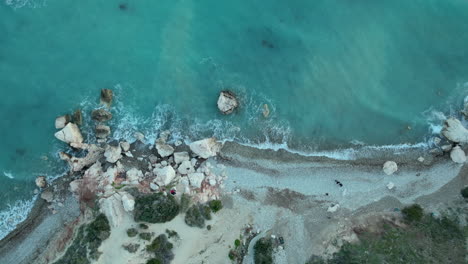 Aerial-perspective-of-a-rocky-beach-with-a-mixture-of-sand-and-boulders,-clear-blue-sea-water-washing-ashore,-with-visible-underwater-rocks-near-the-coastline