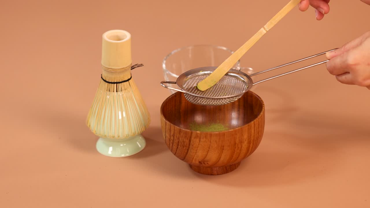 Hand sifts matcha powder through strainer into wooden bowl, minimal background, soft studio lighting