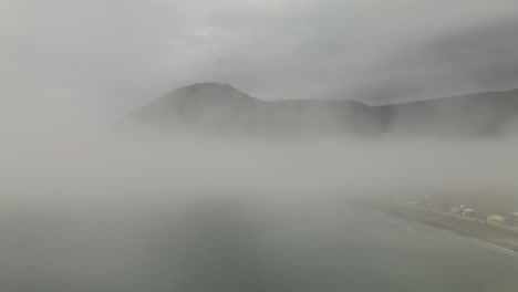 Volando-A-Través-De-La-Niebla-Sombría-Sobre-El-Océano-Por-La-Ciudad-Costera,-El-Pueblo-Chic-chocs,-Para-Revelar-Una-Gran-Montaña-Verde