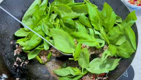 minced meat and fresh basil leaves are stir-fried in a wok under bright lighting, with a spatula mixing the ingredients for a spicy dish