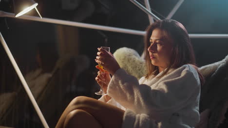 tired woman strokes glass of champagne sitting in armchair. female sits under lamp lighting in room with panoramic windows overlooking night life of nature slow motion