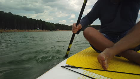 El-Hombre-Rema-Rápido-Mientras-Está-Sentado-En-Paddleboard-Pasando-Por-La-Playa-Del-Lago,-ángulo-Bajo