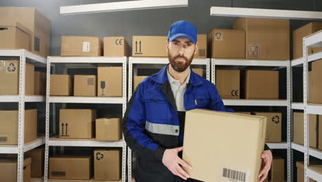Young-Cheerful-Man-In-Uniform-And-Cap-Smiling-Cheerfully-To-Camera-With-Carton-Box-In-Post-Office-Store