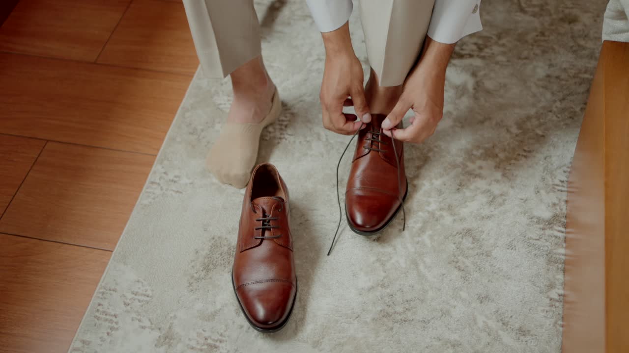 Groom in beige pants tying laces of brown dress shoes on a light rug