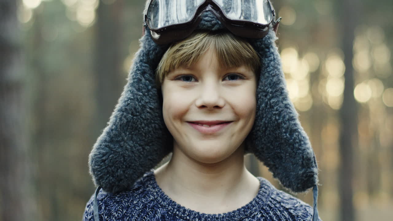 Close-up view of little Caucasian boy dreamer of being a pilot in hat and special glasses smiling at the camera in the forest