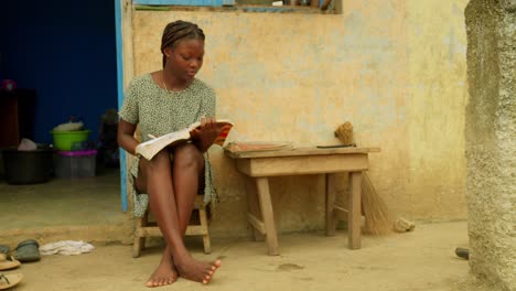 in front of a house, a young woman is studying by reading a book in a village located in kumasi, ghana, in africa