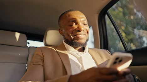 Hombre-De-Negocios-Feliz,-De-Piel-Negra-Y-Barba,-Con-Traje-Marrón,-Charlando-En-Un-Teléfono-Blanco-Y-Conduciendo-Un-Coche-Moderno-Por-La-Ciudad