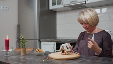 woman standing in modern kitchen, holding an icing bag and toothpick while adjusting decorated gingerbread house cookie on wooden tray