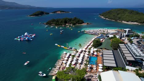 Ksamil-Treasured-Islands:-Tourists-Delighting-in-White-Sand-Beach,-Sun-Chairs,-Umbrellas,-Emerald-Sea,-Aquapark,-Boats,-and-Affordable-Summer-Island-Escapes