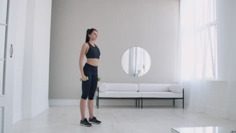 a woman raises her arms with dumbbells doing a shoulder exercise in her light white apartment against a sofa and window.