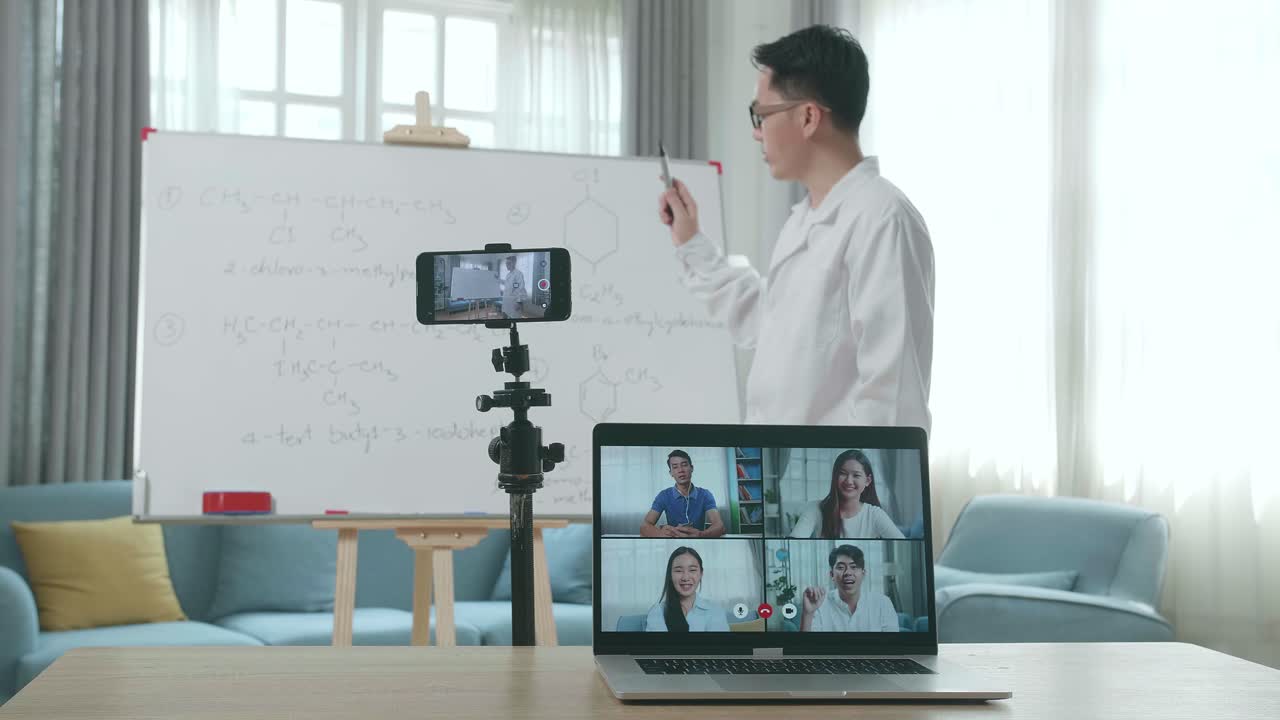 Asian Male Teacher With Glasses Shoots Video By Smartphone Camera And Having Video Call On Laptop While Teaching Chemist At Home