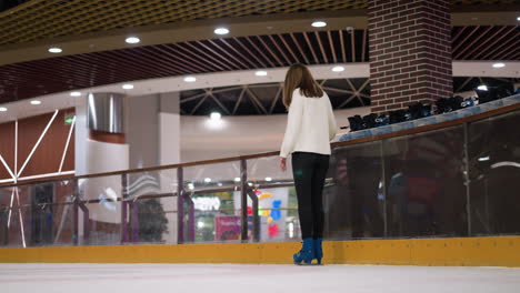 close-up of a lady wearing blue skates, black trousers, and a white top as she skates into the rink, with other persons skating