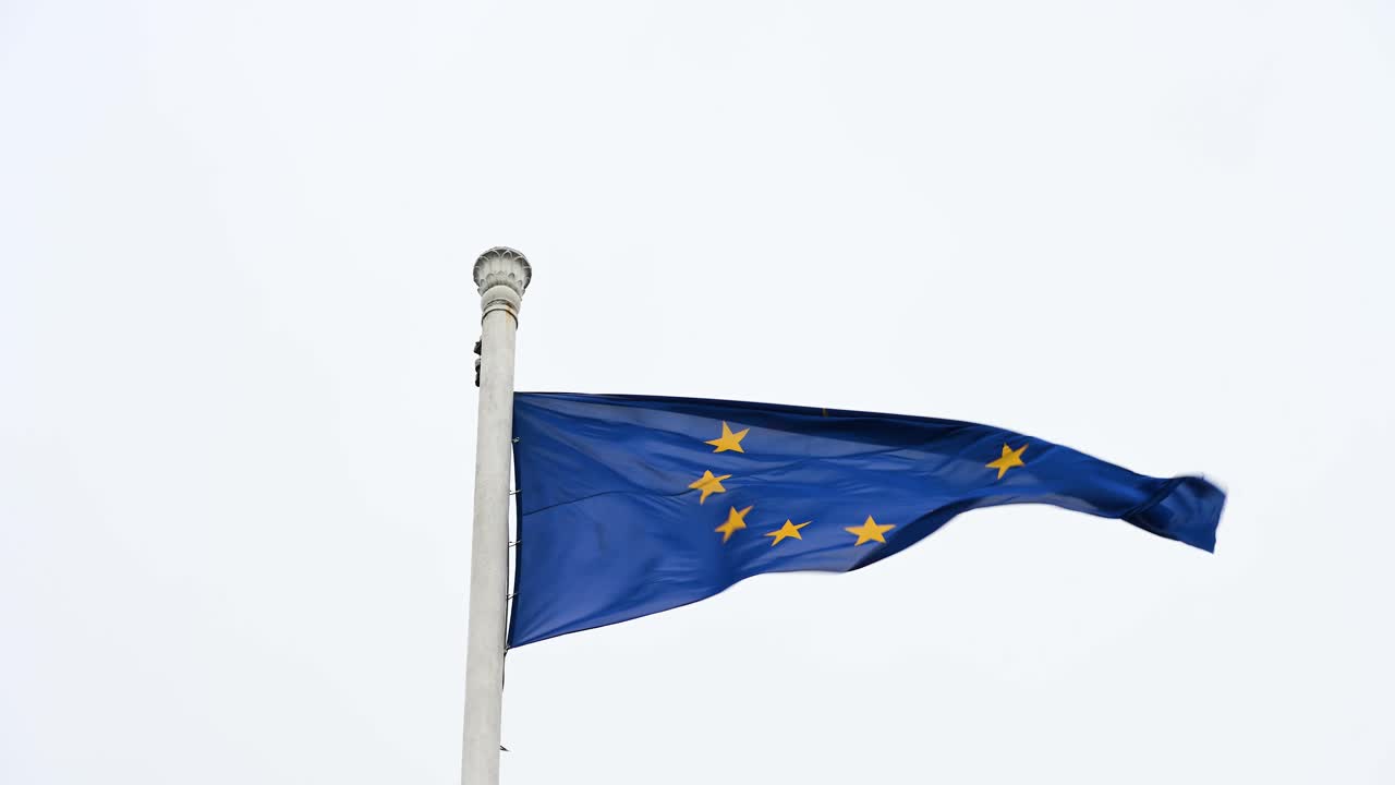 Slow-motion capture of the European Union (EU) flag rippling on a tall post, swaying with the wind on an overcast day, emphasizing the graceful movement of the flag in the elements.