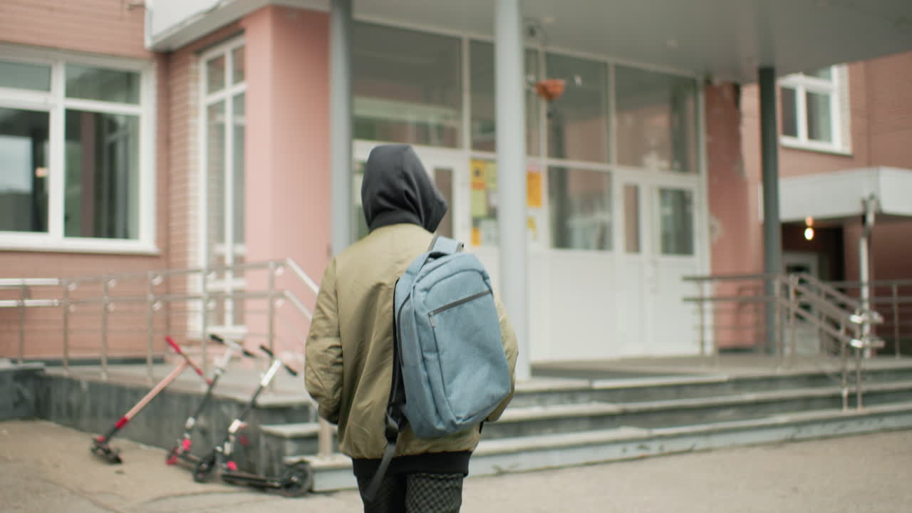 후드 재킷을 입고 배낭을 멘 소년이 강의를 들으러 학교 건물 쪽으로 걸어가고 있어요. 문에 빛이 반사되고 계단 근처에는 스쿠터가 있으며, 흐린 하늘 아래 도시 캠퍼스 환경이 펼쳐져요.