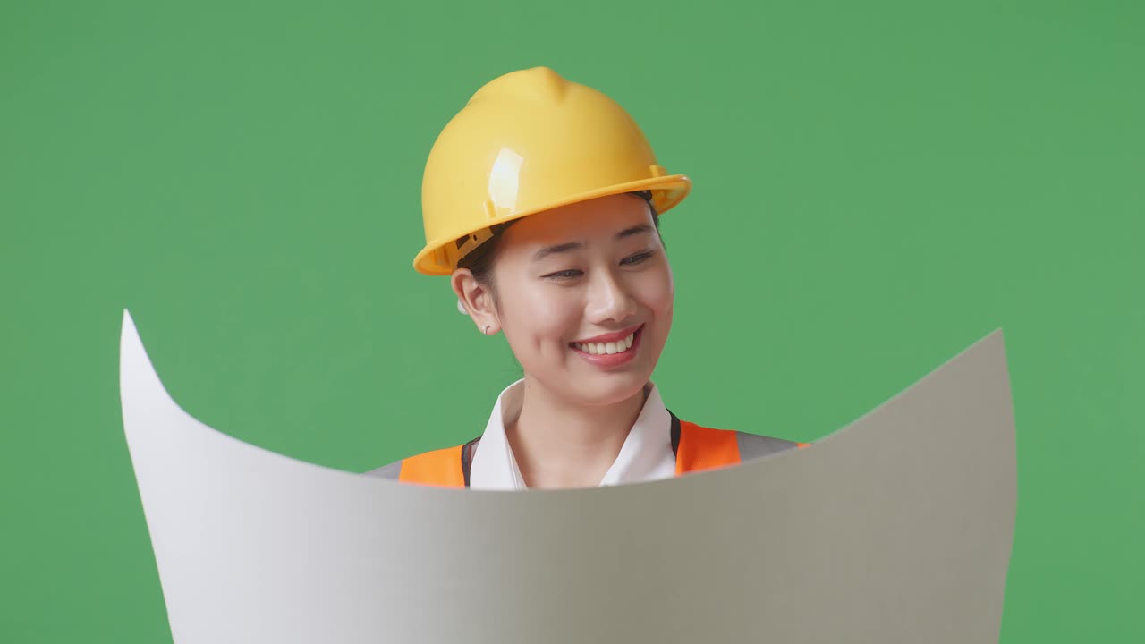 안전 헬을 입은 아시아 여성 엔지니어가 손에 청사진을 들고 녹색 스크린 배경 스튜디오에 서 있는 동안 주변을 둘러보고 있습니다.