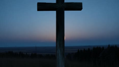 large wooden cross stands on a hilltop, silhouetted against a vibrant sunset in the distance