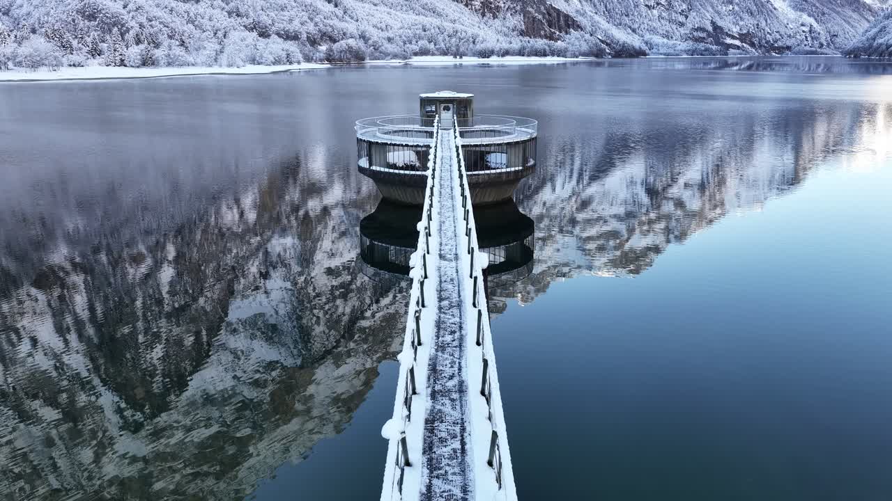 Winter scene of a snow-covered pier leading to a circular reservoir tower on a calm alpine lake in Klontal, Switzerland