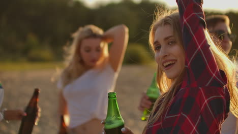 a young female is dancing on the open air party with her friends on the beach. her long blonde hair is flying on the wind. she smiles and enjoys a summertime on the lake coast with beer.